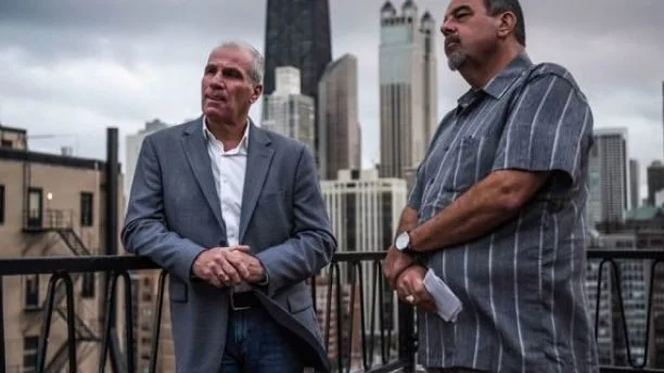 Two investigators on a rooftop with the city skyline behind them